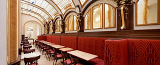 Elegantes Café mit gewölbter, gläserner Decke, roten Polsterbänken und klassischer Inneneinrichtung mit goldenen Wandverzierungen.